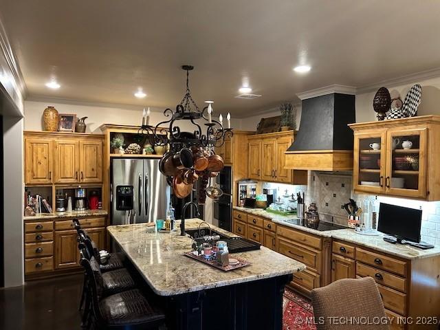 kitchen featuring stainless steel refrigerator with ice dispenser, custom exhaust hood, tasteful backsplash, black electric stovetop, and a kitchen island with sink