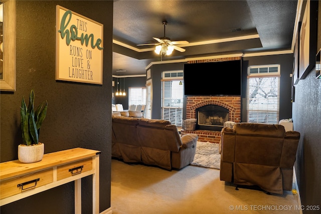 living room with ornamental molding, ceiling fan, a tray ceiling, a brick fireplace, and light carpet