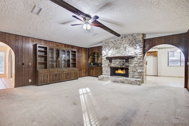 unfurnished living room featuring wooden walls, lofted ceiling with beams, a textured ceiling, light carpet, and a stone fireplace