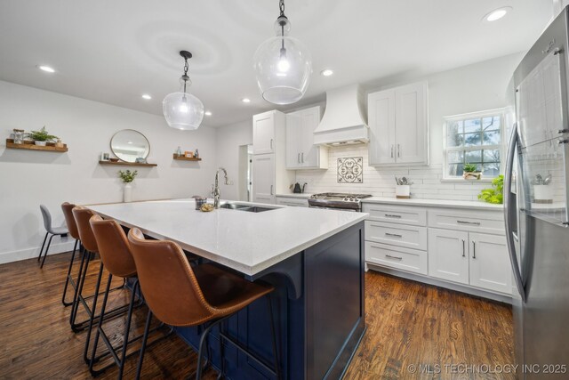 kitchen featuring pendant lighting, a kitchen island with sink, stainless steel appliances, white cabinets, and custom exhaust hood