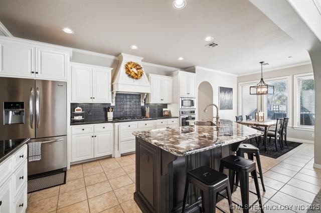 kitchen featuring custom exhaust hood, a center island with sink, light tile patterned floors, appliances with stainless steel finishes, and pendant lighting