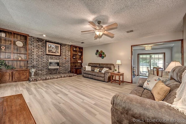 living room featuring built in features, ceiling fan, a brick fireplace, a textured ceiling, and light hardwood / wood-style flooring