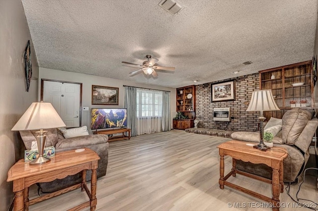 living room featuring built in features, a fireplace, ceiling fan, a textured ceiling, and light wood-type flooring