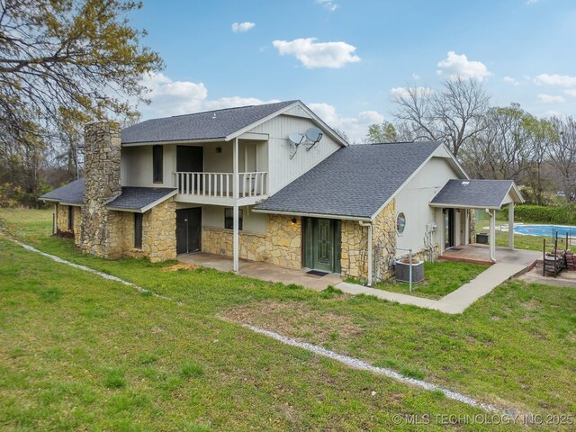 rear view of house with a shingled roof, a patio area, a yard, stone siding, and a chimney