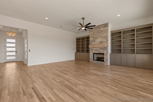 unfurnished living room with a stone fireplace, ceiling fan with notable chandelier, built in features, and light wood-type flooring