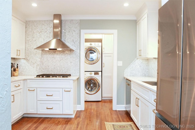 kitchen with stacked washing maching and dryer, white cabinetry, backsplash, stainless steel appliances, and wall chimney range hood