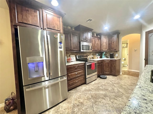 kitchen featuring backsplash, light tile patterned floors, light stone counters, stainless steel appliances, and dark brown cabinets