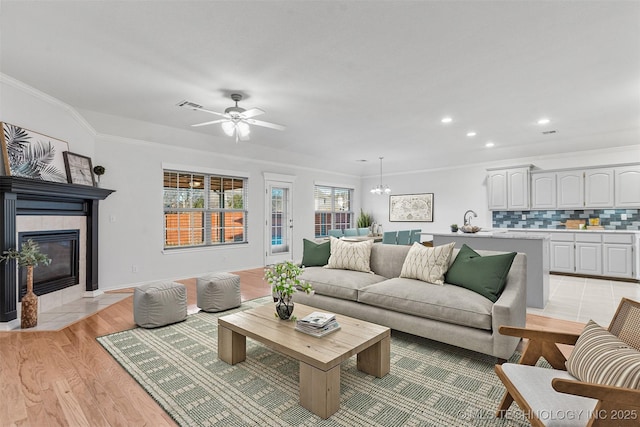 living room featuring ceiling fan with notable chandelier, ornamental molding, a tile fireplace, and light wood-type flooring