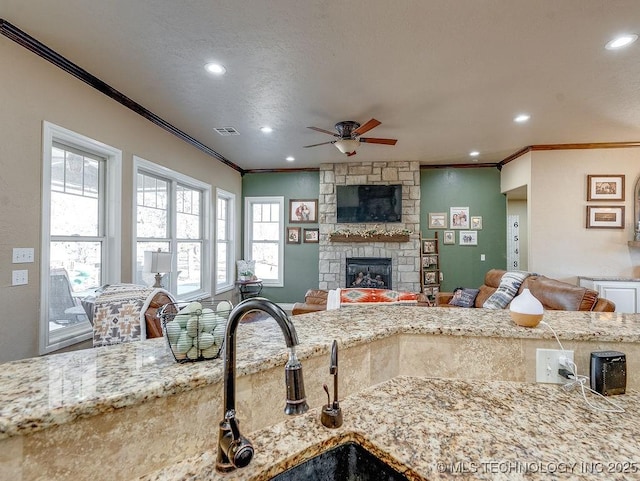 kitchen with ceiling fan, ornamental molding, a stone fireplace, and light stone countertops