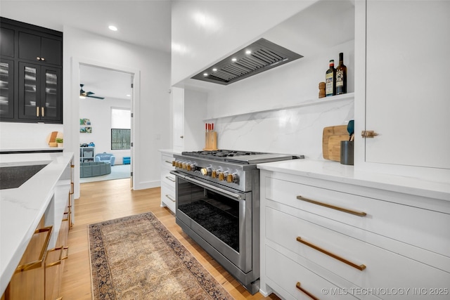 kitchen with wall chimney range hood, stainless steel stove, light stone counters, tasteful backsplash, and light hardwood / wood-style floors