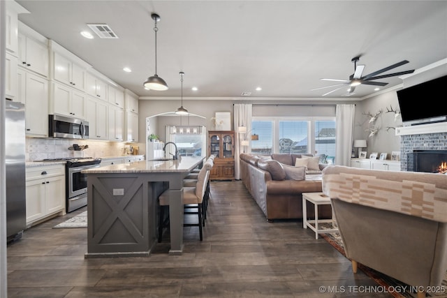 kitchen with stainless steel appliances, light stone counters, an island with sink, white cabinets, and decorative light fixtures