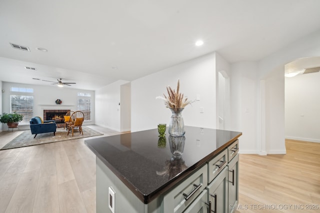 kitchen with a brick fireplace, dark stone countertops, light wood-type flooring, a kitchen island, and ceiling fan