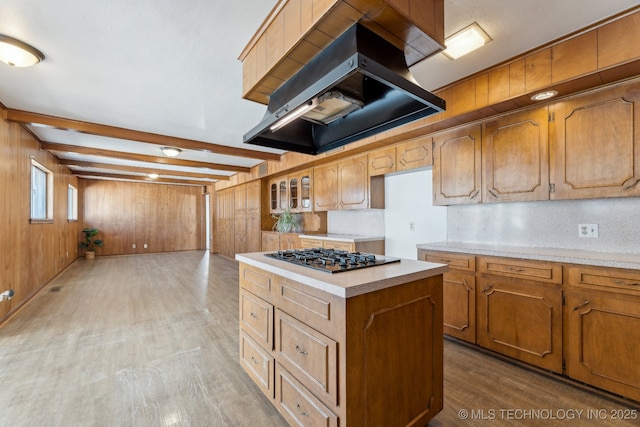 kitchen with beam ceiling, gas stovetop, light countertops, wooden walls, and exhaust hood