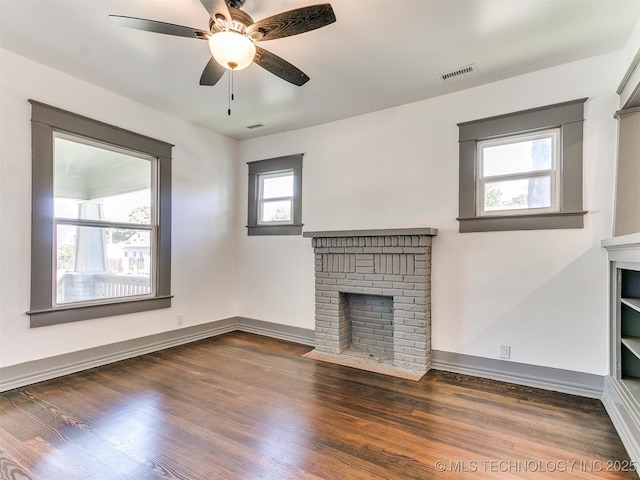 unfurnished living room with a brick fireplace, plenty of natural light, dark wood-type flooring, and ceiling fan