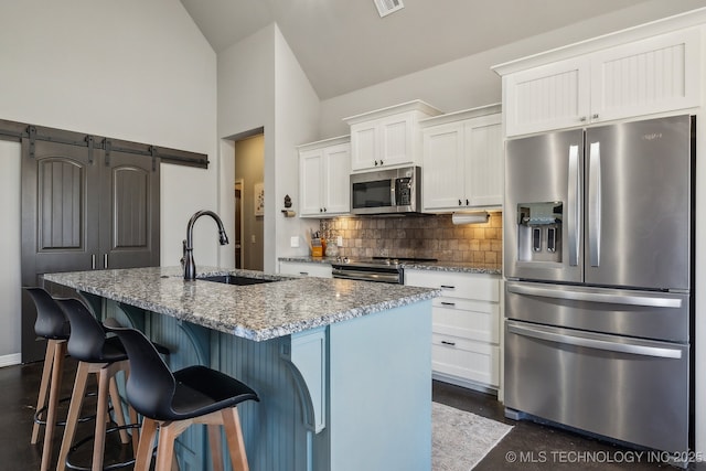 kitchen with appliances with stainless steel finishes, white cabinetry, dark stone countertops, a barn door, and a center island with sink