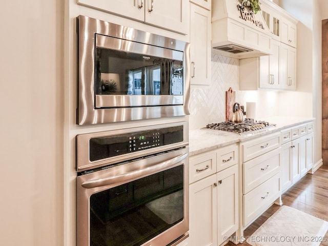 kitchen featuring appliances with stainless steel finishes, backsplash, light stone countertops, white cabinets, and custom exhaust hood