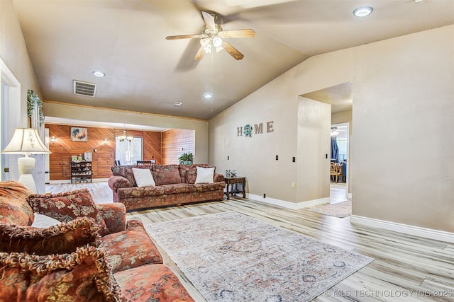 living room featuring ceiling fan with notable chandelier, wooden walls, vaulted ceiling, and light hardwood / wood-style flooring