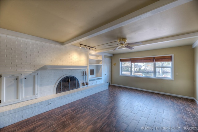 unfurnished living room featuring ceiling fan, a brick fireplace, dark wood-type flooring, track lighting, and beam ceiling