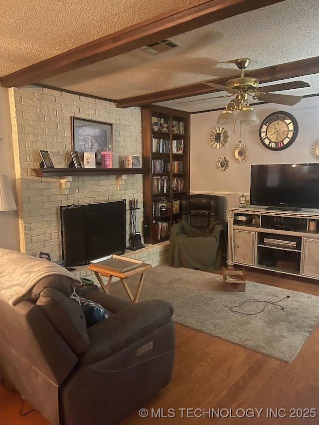 living room featuring a brick fireplace, a textured ceiling, hardwood / wood-style flooring, ceiling fan, and beam ceiling