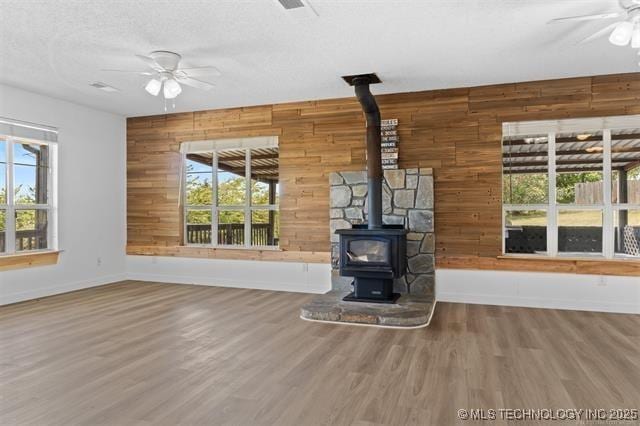 unfurnished living room featuring ceiling fan, wood-type flooring, wooden walls, and a wood stove