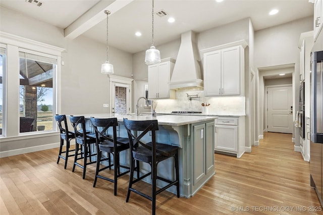 kitchen featuring premium range hood, decorative light fixtures, tasteful backsplash, white cabinets, and a kitchen island with sink