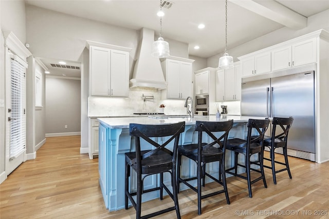 kitchen featuring custom exhaust hood, white cabinetry, built in appliances, hanging light fixtures, and an island with sink