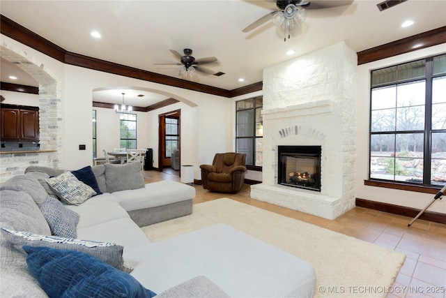 tiled living room with ornamental molding, a stone fireplace, and ceiling fan with notable chandelier