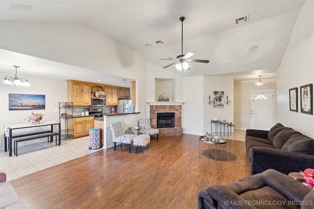 living room with ceiling fan with notable chandelier, a brick fireplace, high vaulted ceiling, and light hardwood / wood-style floors