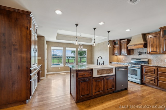 kitchen featuring sink, a kitchen island with sink, stainless steel appliances, custom exhaust hood, and a raised ceiling