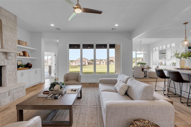 living room with light wood-type flooring, ceiling fan with notable chandelier, built in features, and a fireplace