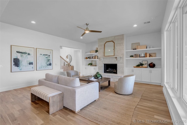 living room featuring built in shelves, a stone fireplace, ceiling fan, and light hardwood / wood-style flooring