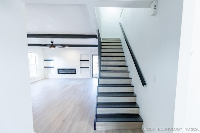 stairs featuring ceiling fan, a glass covered fireplace, beamed ceiling, and wood finished floors