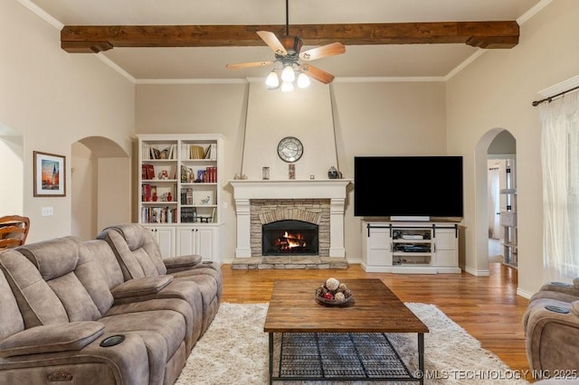 living room with a stone fireplace, crown molding, beamed ceiling, ceiling fan, and light hardwood / wood-style floors