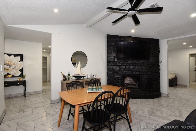 dining room featuring vaulted ceiling with beams, a stone fireplace, a textured ceiling, and ceiling fan