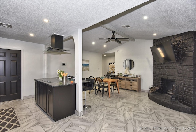 kitchen with ceiling fan, a fireplace, ventilation hood, lofted ceiling with beams, and dark stone counters