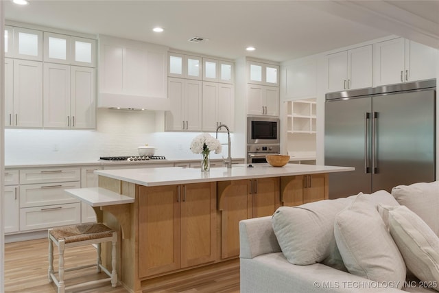 kitchen with built in appliances, sink, white cabinetry, and an island with sink