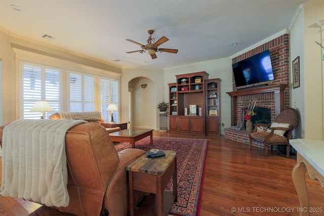 living room featuring crown molding, a brick fireplace, dark wood-type flooring, and ceiling fan