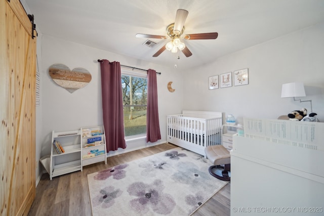 bedroom with a crib, wood-type flooring, a barn door, and ceiling fan