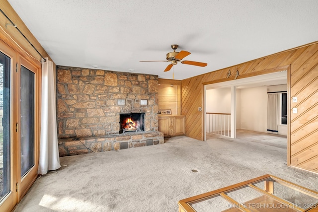 unfurnished living room with a stone fireplace, a textured ceiling, ceiling fan, wooden walls, and light colored carpet