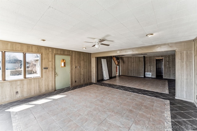 tiled empty room with visible vents, wooden walls, ceiling fan, and stairs