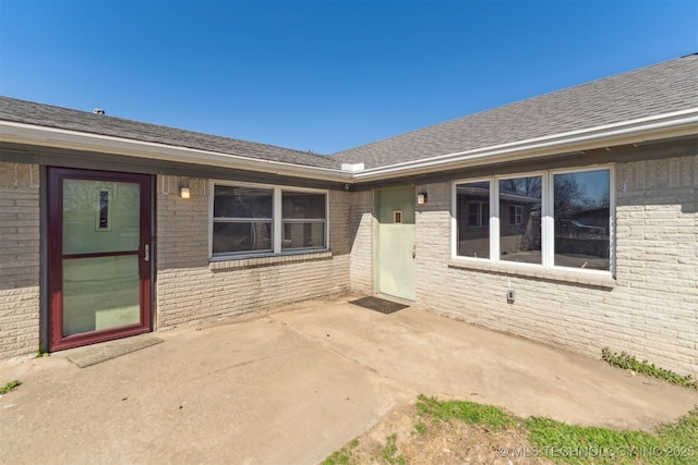 view of exterior entry featuring a patio area, brick siding, and roof with shingles