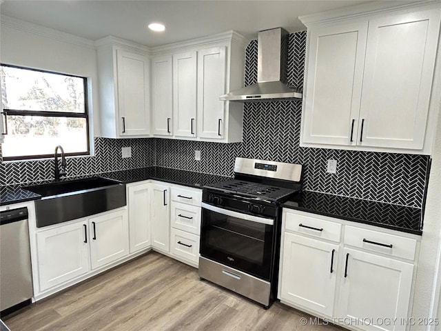 kitchen with sink, appliances with stainless steel finishes, white cabinetry, light hardwood / wood-style floors, and wall chimney exhaust hood