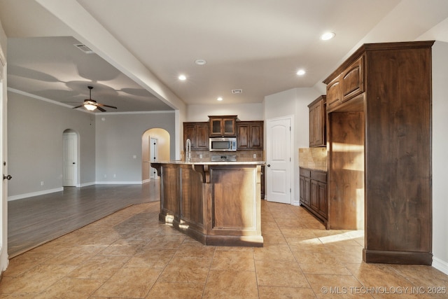 kitchen featuring dark brown cabinetry, ornamental molding, an island with sink, ceiling fan, and decorative backsplash