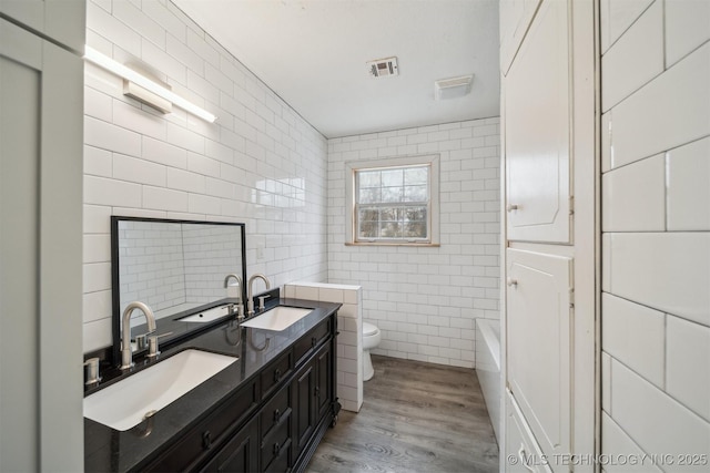 bathroom with tile walls, a tub to relax in, hardwood / wood-style flooring, vanity, and toilet