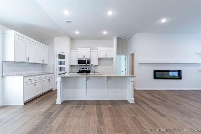 kitchen with light hardwood / wood-style flooring, appliances with stainless steel finishes, a kitchen island with sink, white cabinets, and decorative backsplash