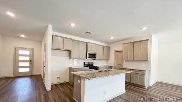 kitchen with dark wood-type flooring, sink, appliances with stainless steel finishes, light stone countertops, and a kitchen island with sink