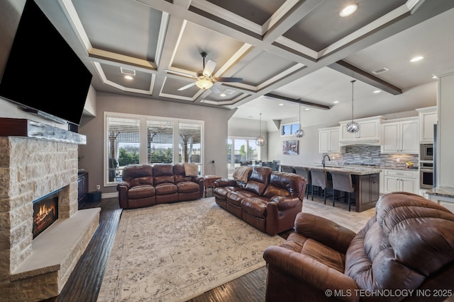 living room featuring a stone fireplace, beamed ceiling, sink, coffered ceiling, and ceiling fan