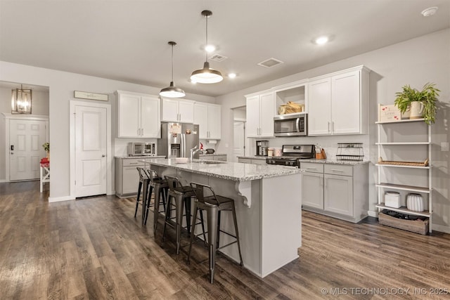 kitchen with a kitchen island with sink, hanging light fixtures, stainless steel appliances, light stone counters, and white cabinets