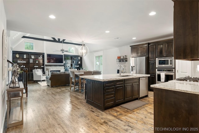 kitchen with appliances with stainless steel finishes, decorative light fixtures, dark brown cabinetry, light stone countertops, and a center island with sink