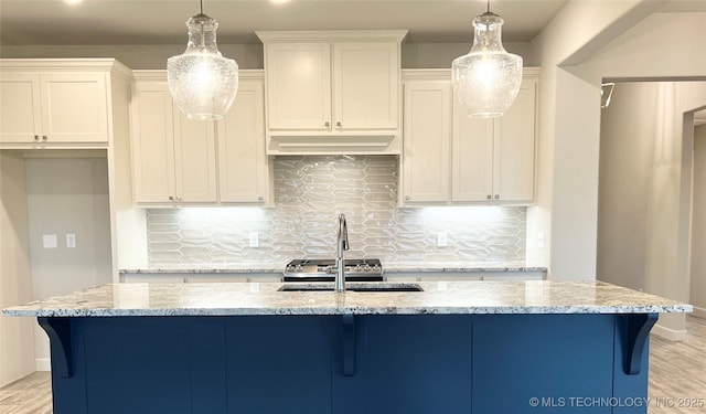 kitchen with light stone counters, pendant lighting, a kitchen island with sink, and white cabinets